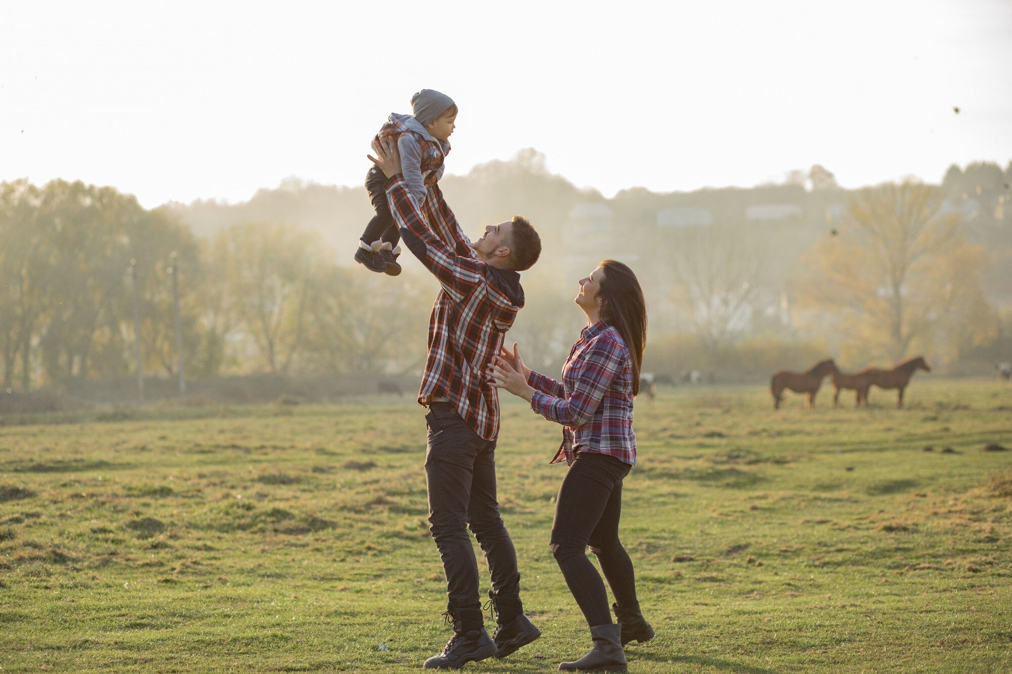 Parents lifting child outdoors in a scenic field with horses grazing in the background.