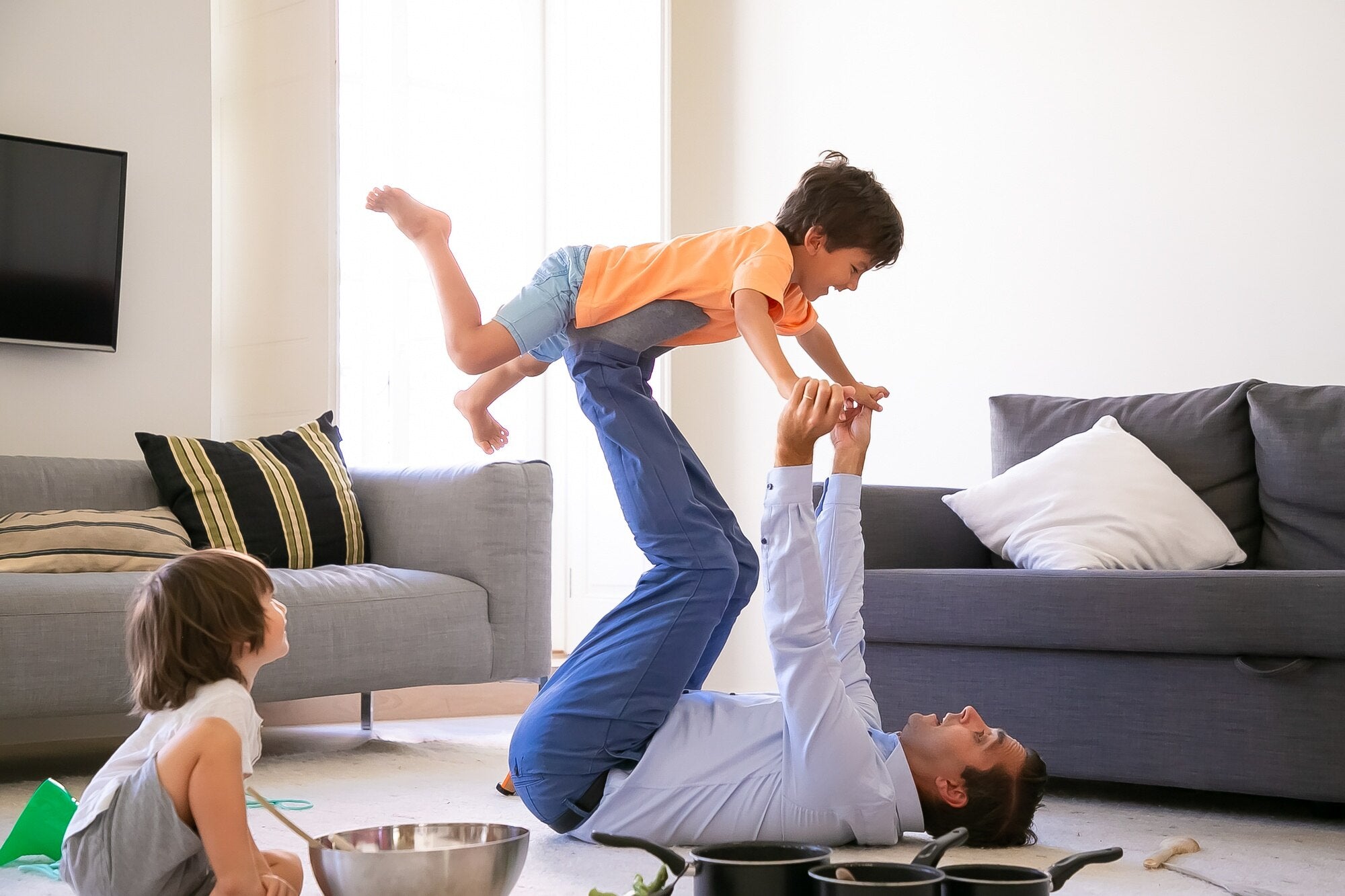 Father playing airplane game with son indoors while daughter watches on the floor.